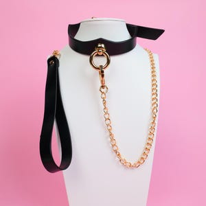 Puede incluir: Un collar de cuero negro con un anillo y una cadena dorados, exhibido en una cabeza de maniquí blanca. El collar tiene una correa a juego con un asa. El fondo es de color rosa liso.