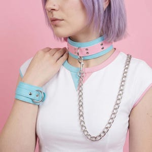 Puede incluir: Un conjunto de collar y puños de colores pastel. El collar es rosa y azul con herrajes plateados y una correa de cadena. El puño a juego es azul claro con una hebilla plateada. El conjunto está diseñado para una estética única.