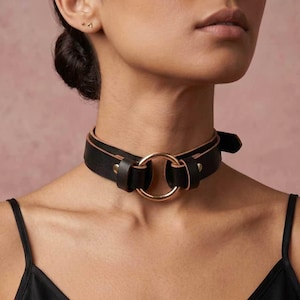 Puede incluir: Un collar gargantilla de cuero negro con un anillo de metal dorado en el centro. El collar tiene un cierre de hebilla y se usa alrededor del cuello. El fondo es de color rosa liso.