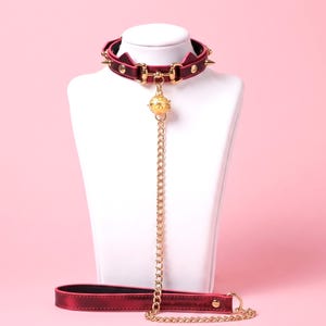 May include: A dark red choker collar featuring gold-tone metal hardware, including small spikes, circular studs, and a central bell charm. A long gold-tone chain connects the collar to a matching dark red leash with a gold-tone metal ring.