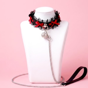 May include: A black lace choker featuring small red ribbon bows and a silver-toned metal ring at the centre. A silver-toned filigree bell hangs from the ring, attached to a long silver-toned metal chain leash with a black leather handle.