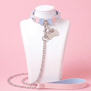 May include: A pastel pink and light blue collar with a bone-shaped centre piece, silver-toned metal hardware, and a matching lead. The collar features two small silver bells and a blank bone-shaped metal tag attached to a central swivel clasp.