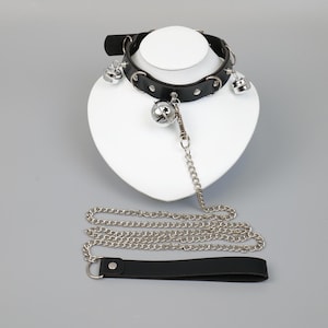 Puede incluir: Collar de cuero negro con campanas plateadas y correa a juego. El collar está tachonado y tiene un cierre de metal. La correa es una cadena plateada con un asa negra.