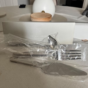 May include: A set of Thomas O'Brien Reed & Barton silverware, including a fork, spoon, and knife, is displayed in its original packaging. The silverware is silver-colored and appears to be stainless steel. A wooden coaster and a white vase with flowers are in the background.