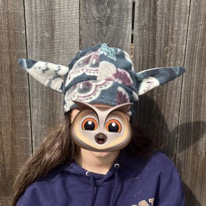 May include: A patterned fleece hat with large, pointed ear-like extensions. The hat features a mix of blue, white, and burgundy floral patterns. The wearer is also wearing a navy blue hoodie with the word "LONDON" printed on it.