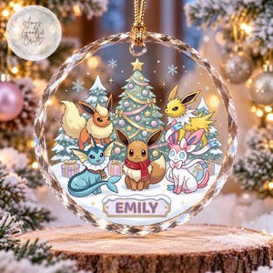 May include: A circular clear glass Christmas ornament featuring a colorful illustration of five Eevee evolution characters around a decorated Christmas tree. The name EMILY is printed in a rectangular banner at the bottom of the ornament.