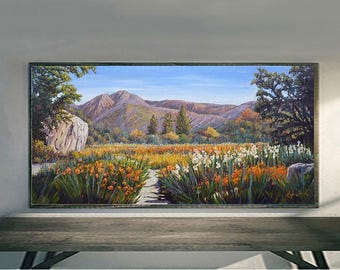 May include: A landscape painting depicting a mountain range under a blue sky. The foreground features a field of orange and white flowers with a stone path leading towards the mountains. The painting is framed in a dark wooden frame.