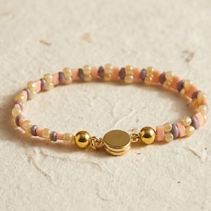May include: A beaded bracelet featuring a repeating pattern of small, round, translucent beige beads, flat peach-colored disc beads, and small, round, dusty purple beads. The bracelet is secured with a polished gold-tone circular magnetic clasp.