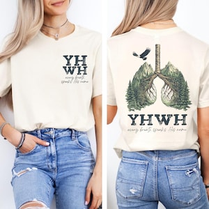 Puede incluir: Camiseta color crema con el texto "YHWH" y "every breath speaks His name" en la parte delantera. La parte trasera presenta un gráfico de pulmones formados por montañas y árboles, con un águila y el mismo texto.