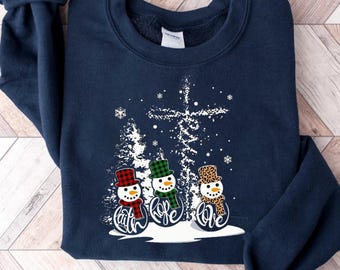 Könnte beinhalten: Marineblaues Sweatshirt mit drei Schneemännern und den Worten "Glaube, Hoffnung, Liebe". Jeder Schneemann trägt einen anderen Hut und Schal. Ein Kreuz und verschneite Bäume im Hintergrund. Ein festliches Winterferien-Design.