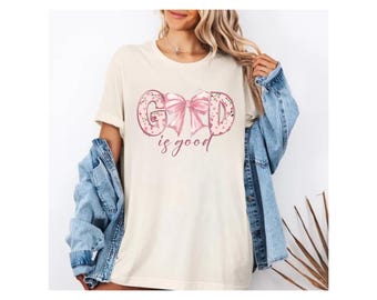 Könnte beinhalten: Cremefarbenes T-Shirt mit dem Schriftzug "GOD is good" in einem rosa, bogenförmigen Design. Die Buchstaben sind mit einem gepunkteten Muster gefüllt. Die Worte "is good" sind in einer Schreibschrift unter dem Bogen geschrieben.