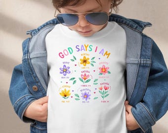 Könnte beinhalten: Weißes T-Shirt mit dem Text "GOD SAYS I AM" und bunten Blumenillustrationen. Das Shirt zeigt Wörter wie "brave", "worthy" und "chosen" um die Blumen herum, mit Bibelversen. Das Kind trägt eine Jeansjacke.