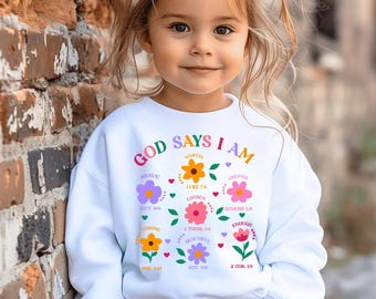 Könnte beinhalten: Ein weißes Sweatshirt mit dem Text "GOD SAYS I AM" in bunten Buchstaben. Das Design umfasst florale Grafiken und Wörter wie "Brave", "Worthy" und "Chosen". Das Sweatshirt wird von einem kleinen Kind getragen.