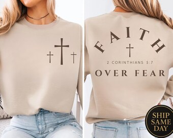 Könnte beinhalten: Beiges Sweatshirt mit drei braunen Kreuzen auf der Vorderseite. Auf der Rückseite des Sweatshirts steht das Wort "FAITH" über einem Kreuz, mit "2 Corinthians 5:7" und "OVER FEAR" darunter.