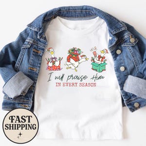 Puede incluir: Una camiseta blanca debajo de una chaqueta vaquera azul. La camiseta presenta un gráfico de tres ocas con gorros de Papá Noel, rodeadas de regalos y luces. El texto dice, I will praise Him IN EVERY SEASON en letras negras y rojas.