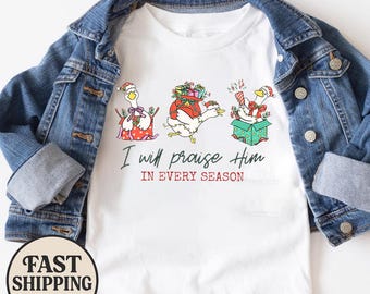 Könnte beinhalten: Ein weißes T-Shirt unter einer blauen Jeansjacke. Das Shirt zeigt eine Grafik von drei Gänsen mit Weihnachtsmützen, umgeben von Geschenken und Lichtern. Der Text lautet, I will praise Him IN EVERY SEASON in schwarzer und roter Schrift.