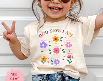 Könnte beinhalten: Ein naturfarbenes T-Shirt mit dem Text "GOD SAYS I AM" und bunten Blumengrafiken. Die Blumen haben Wörter wie "Brave", "Chosen" und "Beautiful". Das T-Shirt wird von einem Kind getragen.
