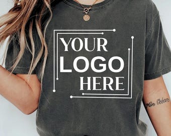 Könnte beinhalten: Dunkelgraues T-Shirt mit dem Text "YOUR LOGO HERE" in einem weißen, umrandeten Feld. Das T-Shirt hat einen Rundhalsausschnitt und scheint aus einem weichen Material zu sein. Das T-Shirt ist einfarbig und hat kurze Ärmel.