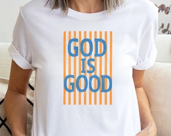 Könnte beinhalten: Weißes T-Shirt mit dem Text "GOD IS GOOD" in blauen Buchstaben, vor einem Hintergrund aus vertikalen orangefarbenen Streifen. Das Shirt hat kurze Ärmel und einen Rundhalsausschnitt.