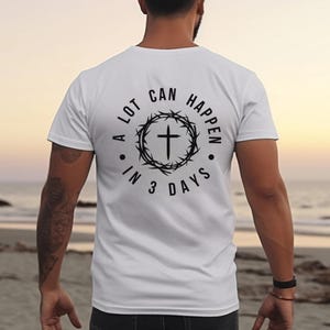 Puede incluir: Camiseta blanca con un gráfico negro en la espalda. El gráfico presenta una cruz dentro de un círculo de espinas, con el texto "A LOT CAN HAPPEN IN 3 DAYS" que rodea el diseño. La camiseta es sencilla y lisa.