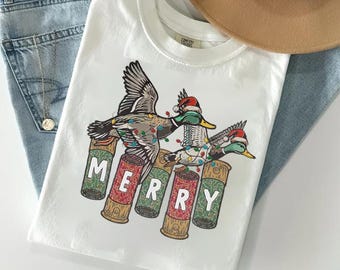Könnte beinhalten: Ein weißes T-Shirt mit einer Grafik von zwei Stockenten mit Weihnachtsmützen, die sich in bunten Lichterketten verfangen haben und über fünf vertikal angeordneten Schrotpatronen fliegen. Das Wort MERRY ist auf die Patronen gedruckt.