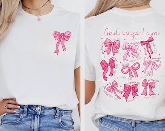 Könnte beinhalten: Weißes T-Shirt mit einer rosa Schleifen-Grafik auf der Vorder- und Rückseite. Auf der Rückseite des Shirts steht der Text "Gott sagt, ich bin" und mehrere rosa Schleifengrafiken mit Wörtern wie "geschätzt" und "wertvoll".