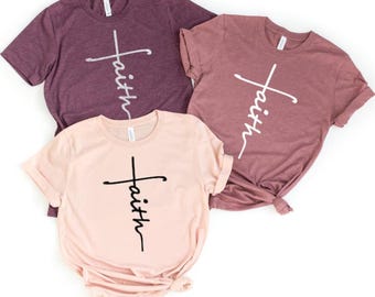 Könnte beinhalten: Drei kurzärmelige T-Shirts in Rosa- und Lilatönen. Jedes Shirt hat ein Kreuzdesign mit dem Wort "faith" vertikal geschrieben. Die Shirts sind aus weichem Material und für Freizeitkleidung geeignet.
