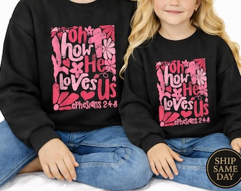 Könnte beinhalten: Schwarze Sweatshirts mit dem Text "Oh how He loves Us" in Rosa und Weiß, mit Blumen- und Herzmotiven. Der Text "Epheser 2:4-8" ist ebenfalls enthalten. Die Sweatshirts werden von zwei Personen getragen.