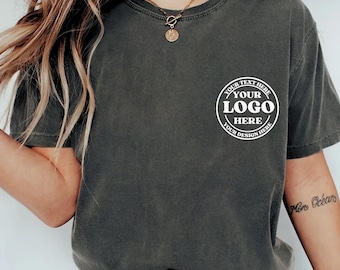 Könnte beinhalten: Dunkelgraues T-Shirt mit einem weißen kreisförmigen Logo auf der linken Brust. Das Logo lautet "YOUR TEXT HERE", "YOUR LOGO HERE" und "YOUR DESIGN HERE" in einem kreisförmigen Design. Das Shirt hat einen Rundhalsausschnitt und kurze Ärmel.