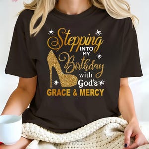 Puede incluir: Una camiseta de manga corta color marrón oscuro con un gráfico de zapato de tacón alto con purpurina dorada y el texto "Stepping INTO MY Birthday with God's GRACE & MERCY" en fuentes cursivas y de bloque, con pequeños iconos de estrellas.