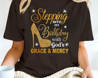 Könnte beinhalten: Ein dunkelbraunes Kurzarm-T-Shirt mit einer Grafik eines goldglitzernden High-Heel-Schuhs und dem Text "Stepping INTO MY Birthday with God's GRACE & MERCY" in einer Mischung aus Schreib- und Blockschrift, akzentuiert durch kleine weiße Sterne.