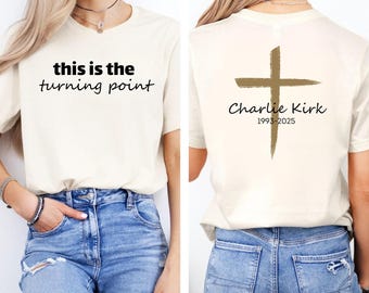 Könnte beinhalten: Cremefarbenes T-Shirt mit dem Text "this is the turning point" auf der Vorderseite. Auf der Rückseite des Shirts befindet sich ein goldfarbenes Kreuz und der Name "Charlie Kirk" mit den Jahren 1993-2025.