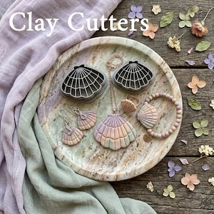 Op de afbeelding: Een verzameling kleisnijders en sieraden met schelpmotieven, waaronder oorbellen, een ketting, een ring en een armband. De items zijn in pastelkleuren en gerangschikt op een marmeren dienblad. De tekst "Clay Cutters" staat bovenaan.