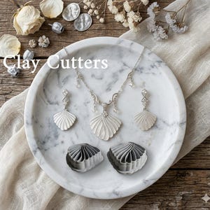 Op de afbeelding: Een zilveren schelpvormige ketting en oorbellen set met heldere kristallen accenten, gepresenteerd op een marmeren dienblad. Er zijn ook twee schelpvormige kleisnijders aanwezig. De tekst "Clay Cutters" staat in de afbeelding.