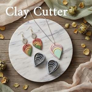 Può includere: Una collezione di gioielli in argilla polimerica fatti a mano e taglierine per argilla. I gioielli includono orecchini e una collana con ciondoli a forma di foglia nei toni del rosa, verde e giallo. L'immagine mostra anche taglierine per argilla grigie a forma di goccia. Il testo "Clay Cutter" è in alto.