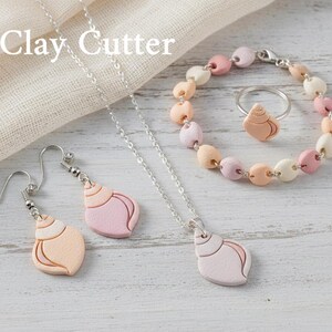 Può includere: Un set di gioielli con un design a conchiglia. Il set include orecchini, una collana, un braccialetto e un anello. I gioielli sono nelle tonalità del rosa, pesca e bianco. Il testo "Clay Cutter" è visibile nell'immagine.