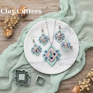 Op de afbeelding: Een sieradenset en kleisnijders worden gepresenteerd op een marmeren bord en een houten oppervlak. De sieraden omvatten een ketting, oorbellen en een ring, allemaal met een geometrisch ontwerp met blauwe, roze en bloemige accenten. De tekst "Clay Cutters" is zichtbaar.
