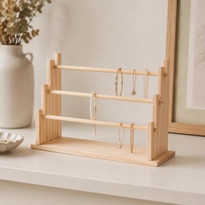 May include: A three-tiered light wood jewelry display stand featuring horizontal rods for hanging necklaces and bracelets. The structure is crafted from natural wood with a visible grain, providing a stable base and vertical supports to organize jewelry items.