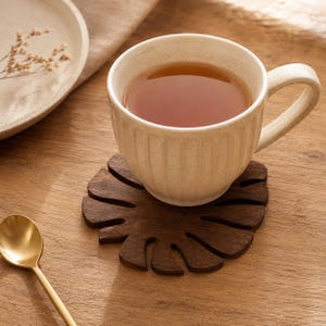 Peut inclure: Une tasse en céramique de couleur crème avec des détails nervurés verticaux remplie de thé, reposant sur un dessous de verre en bois brun foncé en forme de feuille de monstera. La tasse présente une anse incurvée et une finition lisse.