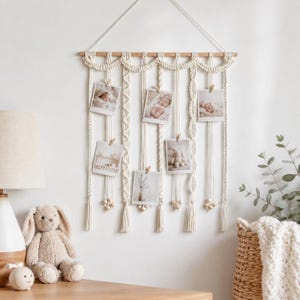 May include: A cream-colored macrame wall hanging suspended from a wooden dowel, featuring intricate braided patterns and vertical cords. Five small rectangular photographs are attached to the cords with wooden clips, each displaying images of infants.