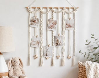 May include: A cream-colored macrame wall hanging suspended from a wooden dowel, featuring intricate braided patterns and vertical cords. Five small rectangular photographs are attached to the cords with wooden clips, each displaying images of infants.