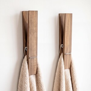 May include: Two wooden clothespin-shaped wall hooks with towels hanging from them. The hooks are made of dark brown wood and have a silver-colored metal spring mechanism. The towels are a light beige color, and the background is a plain white wall.