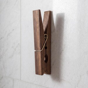 May include: A large, wooden clothespin-shaped wall hook. The hook is made of dark brown wood with a brass-colored metal spring. The clothespin is mounted on a white marble-like wall, creating a decorative and functional item.