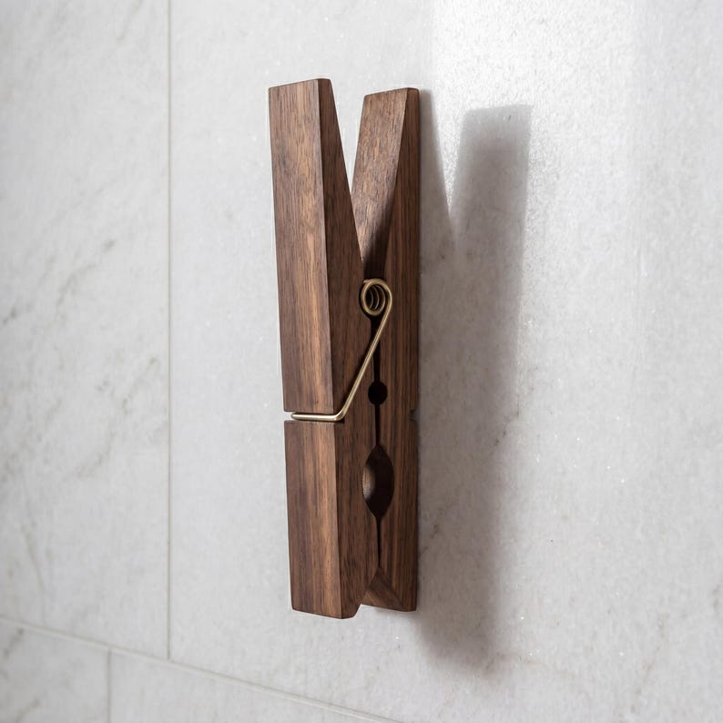 May include: A large, wooden clothespin-shaped wall hook. The hook is made of dark brown wood with a brass-colored metal spring. The clothespin is mounted on a white marble-like wall, creating a decorative and functional item.