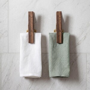 May include: Two wooden towel holders with brass accents are mounted on a marble tile wall. Each holder features a large, dark wood clothespin design, holding a folded towel. One towel is white, and the other is a muted green.
