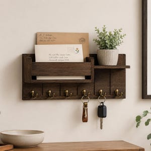 May include: A dark brown wooden wall-mounted organiser featuring a mail slot, a small shelf holding a potted green plant, and six brass-coloured metal hooks. Two sets of keys hang from the hooks, and two envelopes are tucked into the mail slot.