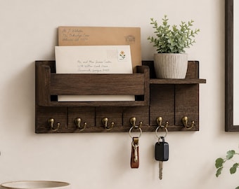 May include: A dark brown wooden wall-mounted organizer featuring a mail slot, a small shelf holding a potted green plant, and six brass-colored metal hooks. Two sets of keys hang from the hooks, and two envelopes are tucked into the mail slot.
