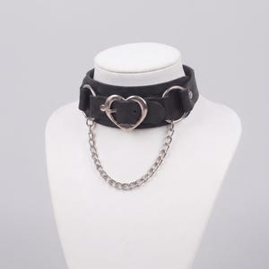 May include: A black choker necklace featuring a central silver-toned heart-shaped buckle, two silver-toned metal rings on the sides, and a silver-toned chain that connects the two rings, draping below the heart buckle in a curved shape.