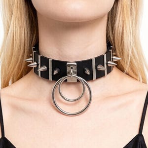 Puede incluir: Un collar gargantilla negro con detalles metálicos plateados y púas. El collar tiene un colgante de doble anillo. Un accesorio llamativo que se lleva alrededor del cuello.