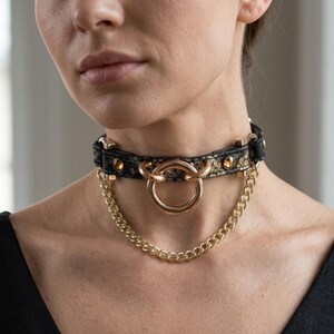 May include: A black choker necklace featuring a textured snakeskin pattern, accented with gold-tone hardware including a central circular ring, two small amber-colored studs, and a delicate gold-tone chain draped beneath the main collar band.